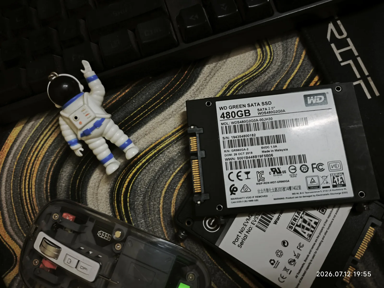 A WD Green 480GB SATA SSD pulled out of the desktop, sitting next to a mechanical keyboard, a small astronaut keychain, and a partially disassembled remote, mid desk teardown.